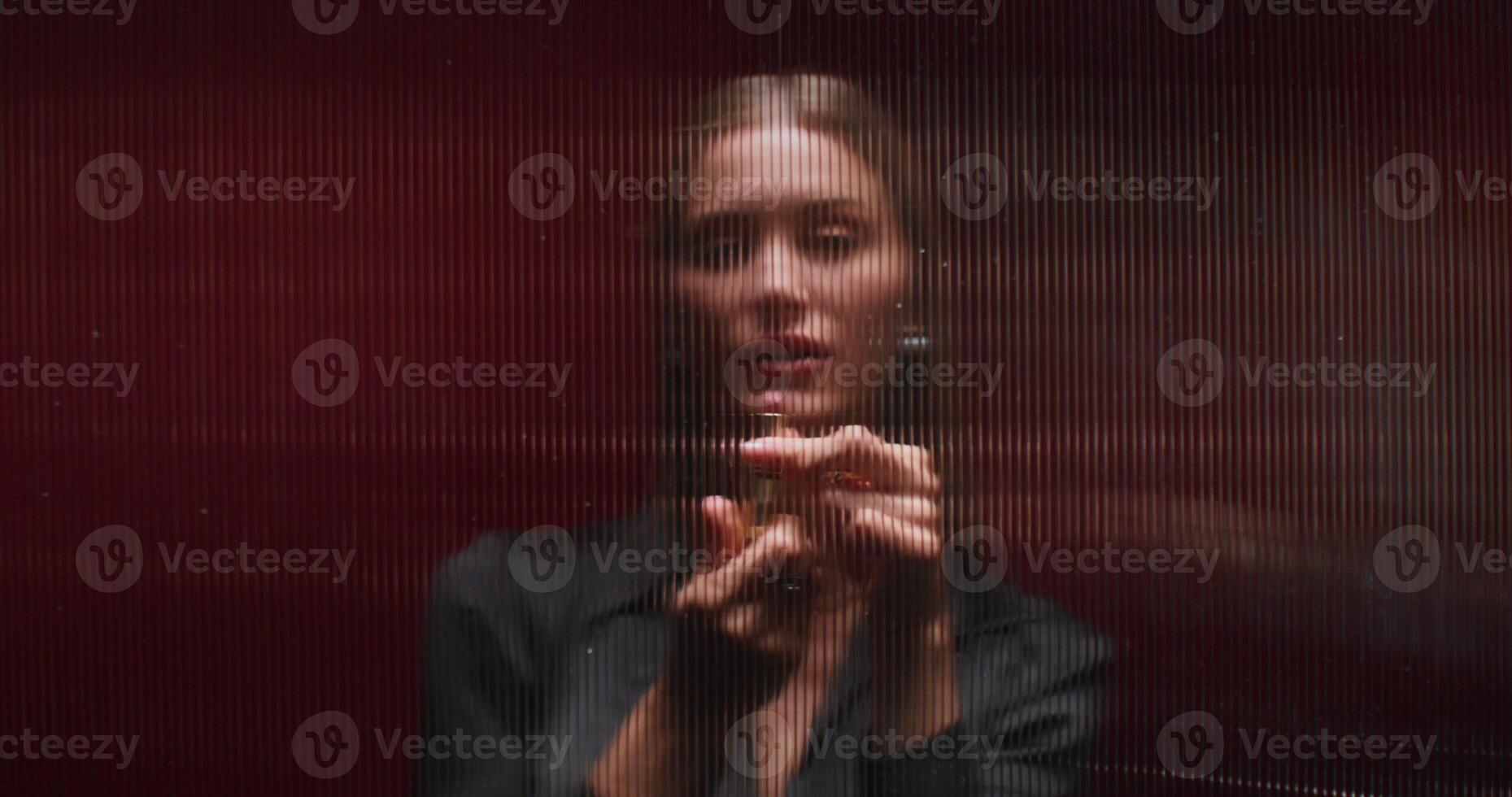 A woman paints her lips with lipstick standing behind a striped glass. She looks at the camera with a serious look. Studio photo