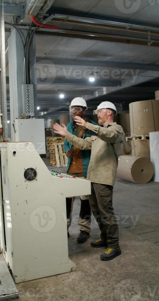 Two men in protective helmets and glasses are talking standing near the machine control panel at a paper mill photo