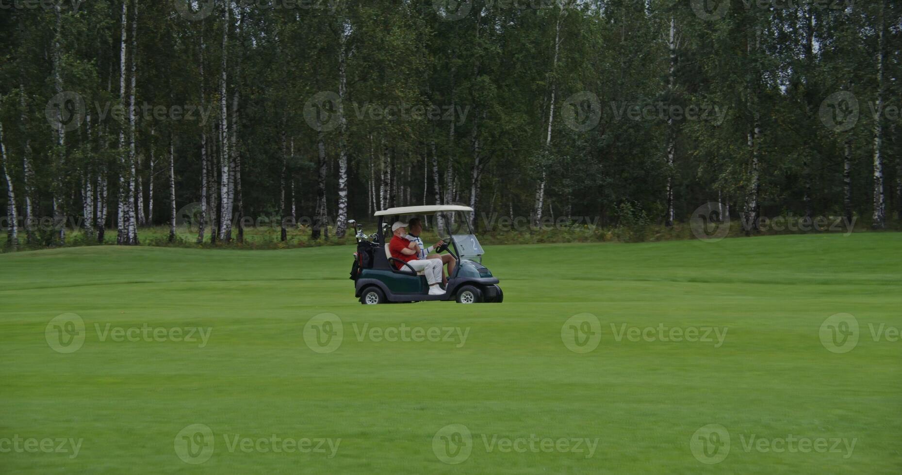 Side view of a golf cart driving through a golf course with two men and equipment. Well-groomed golf course photo