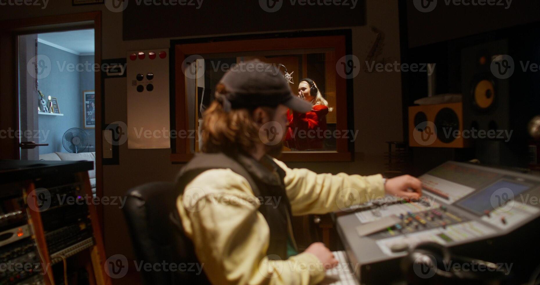 Working in a recording studio. The sound engineer uses an audio mixer. The singer sings into the microphone behind the glass photo