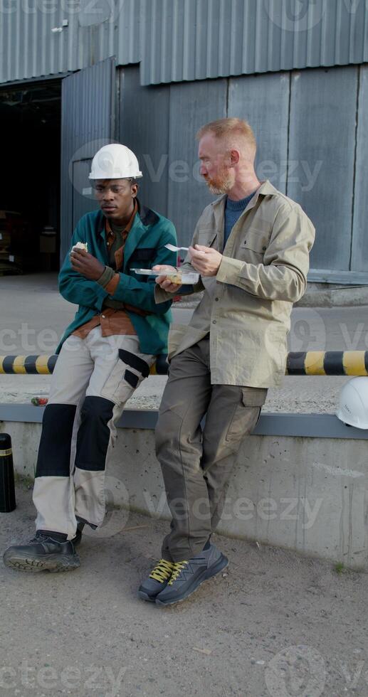 Two workers are eating while sitting on a ramp near the factory. Lunch break at the factory. They're talking. photo