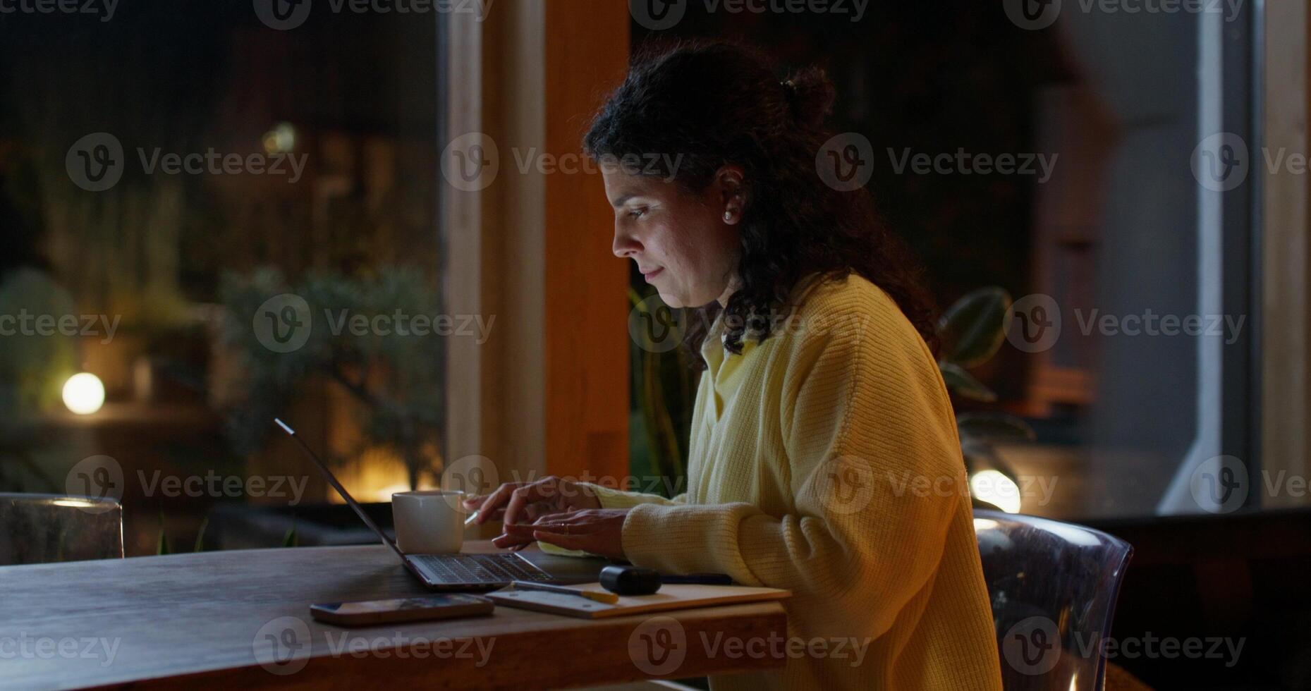 The woman works from home at night. She uses a laptop and writes in a notebook while sitting at her desk. photo