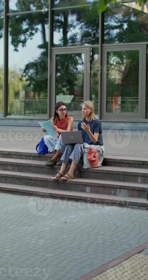 Two women are using a laptop while sitting on the building steps. photo