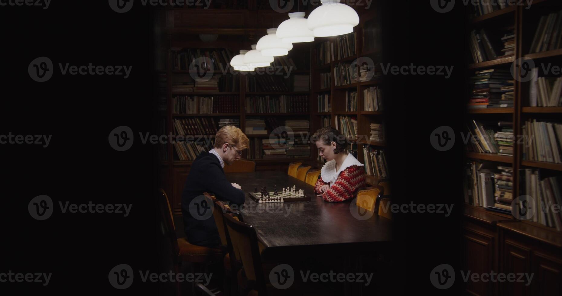 Young man and woman of European appearance playing a chess game, thinking intently, sitting opposite each other at a table in the library photo