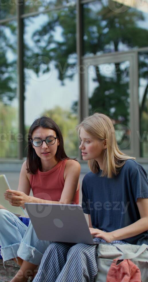 Twowomen are using a laptop while sitting on the building steps. They are talking and smiling. Summer day photo