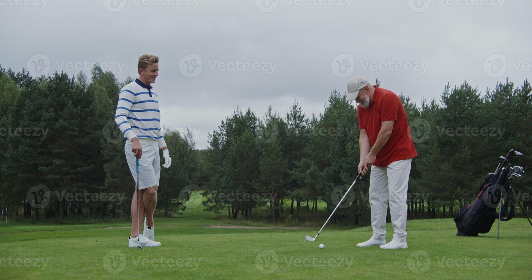Father and son play golf together, talking and laughing. A young handsome man of European appearance looks as an elderly gray-haired man with a beard is hitting ball with a golf club. photo