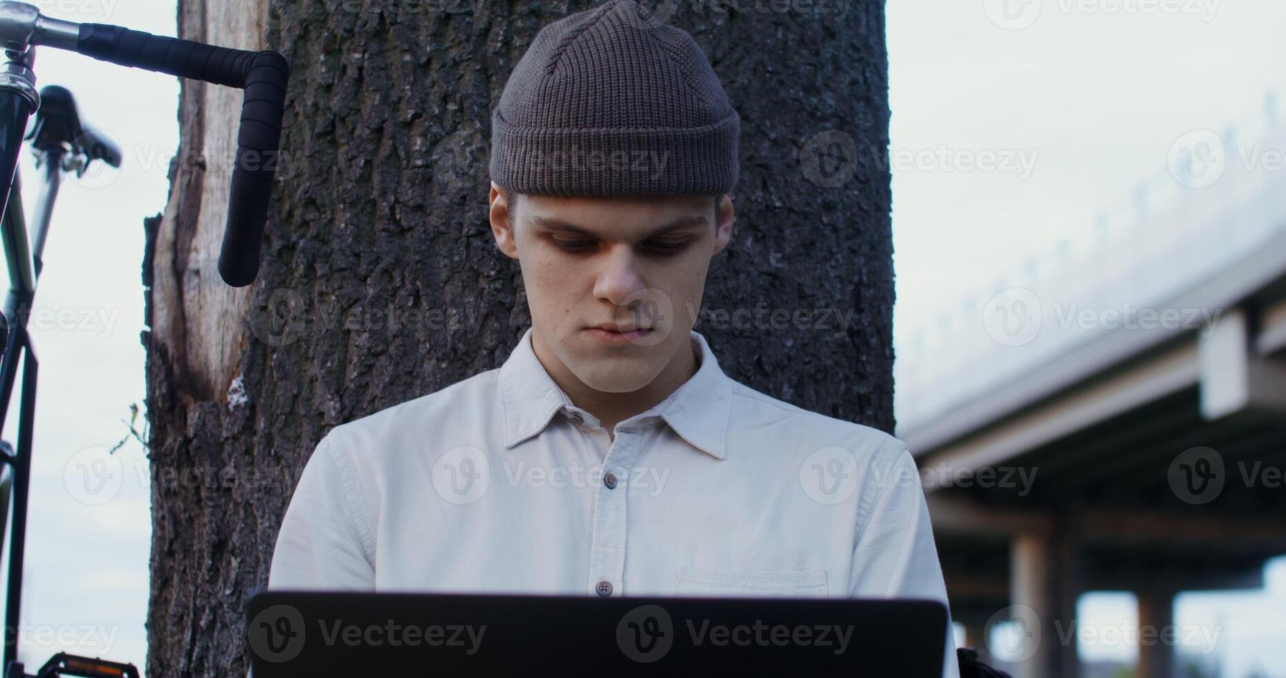 young man of European appearance, in a knitted hat, uses a laptop, sitting on the ground under a tree in the open air. Bicycle Stands next to a man photo
