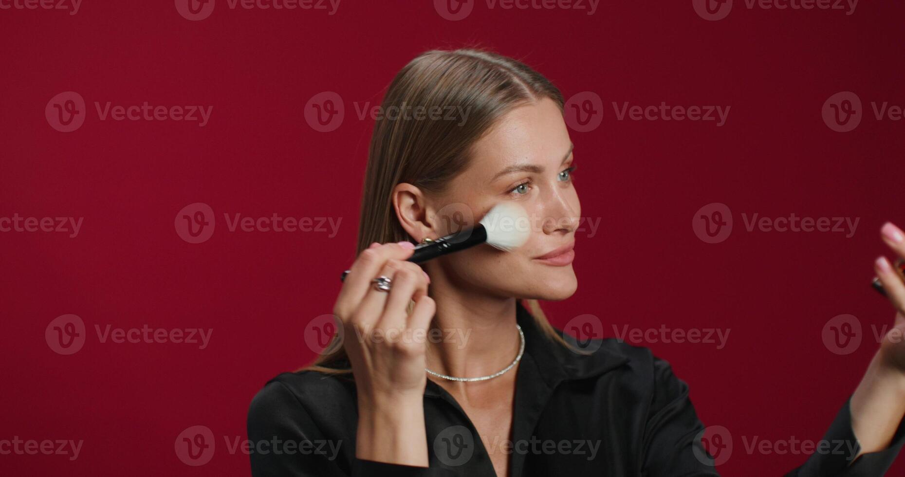 A woman in formal clothes applies blush to her face with a brush. She smiles at the camera while standing in the studio photo
