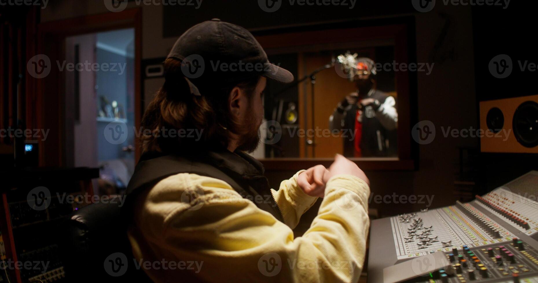 The rapper is recording song in the studio. The sound engineer controls the recording process sitting in the control room photo