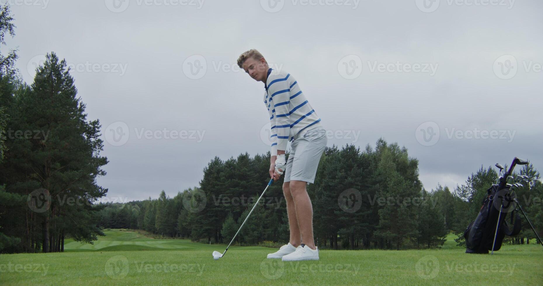 Handsome blond man playing golf on a large field alone. Ball hitting with a golf club, dynamic action. Stylish golf apparel and equipment photo