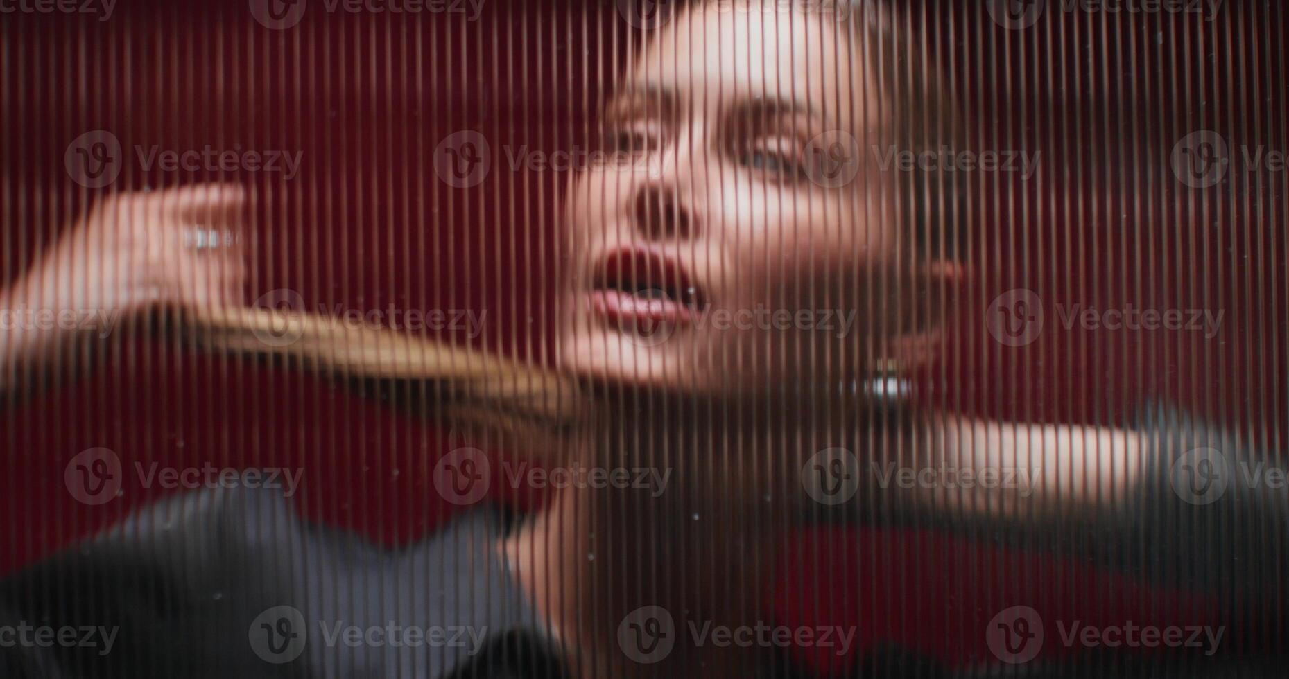 A woman looks at the camera standing behind a striped glass. She's posing in the studio. Close-up photo