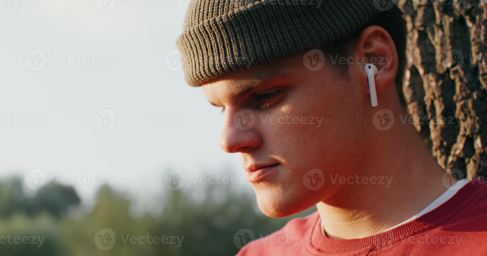 young man of European appearance in a knitted hat and with wireless headphones in his ears sits on the ground under a tree and looks down intently, close-up, only his face is in the frame photo