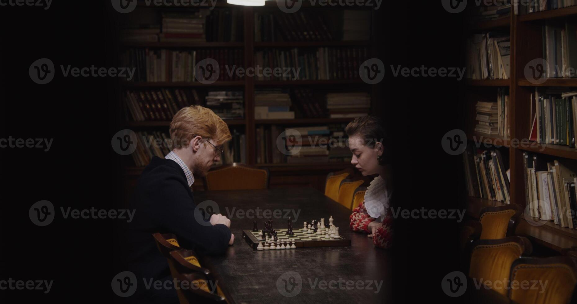 Young Caucasian couple playing chess at a table in a library, both deep in thought. photo