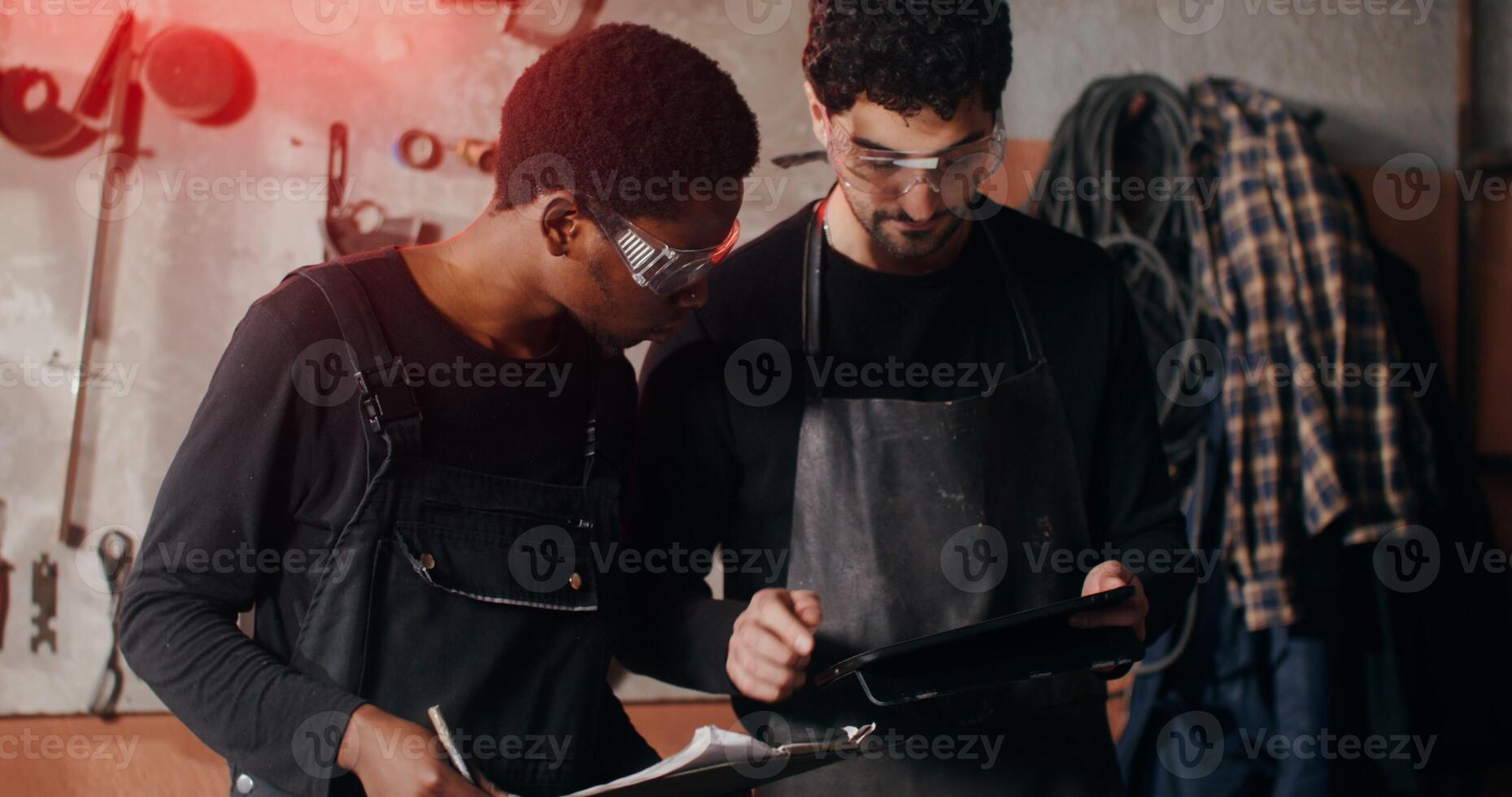 Two blacksmiths of different nationalities are thinking about a sketch of a future product, working together and using a tablet to search for ideas, in workshop full of tools photo