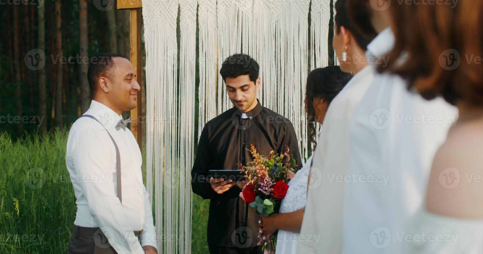 Wedding of an African American couple in a pine forest. A young priest with a tablet. Decorated arch and guests photo