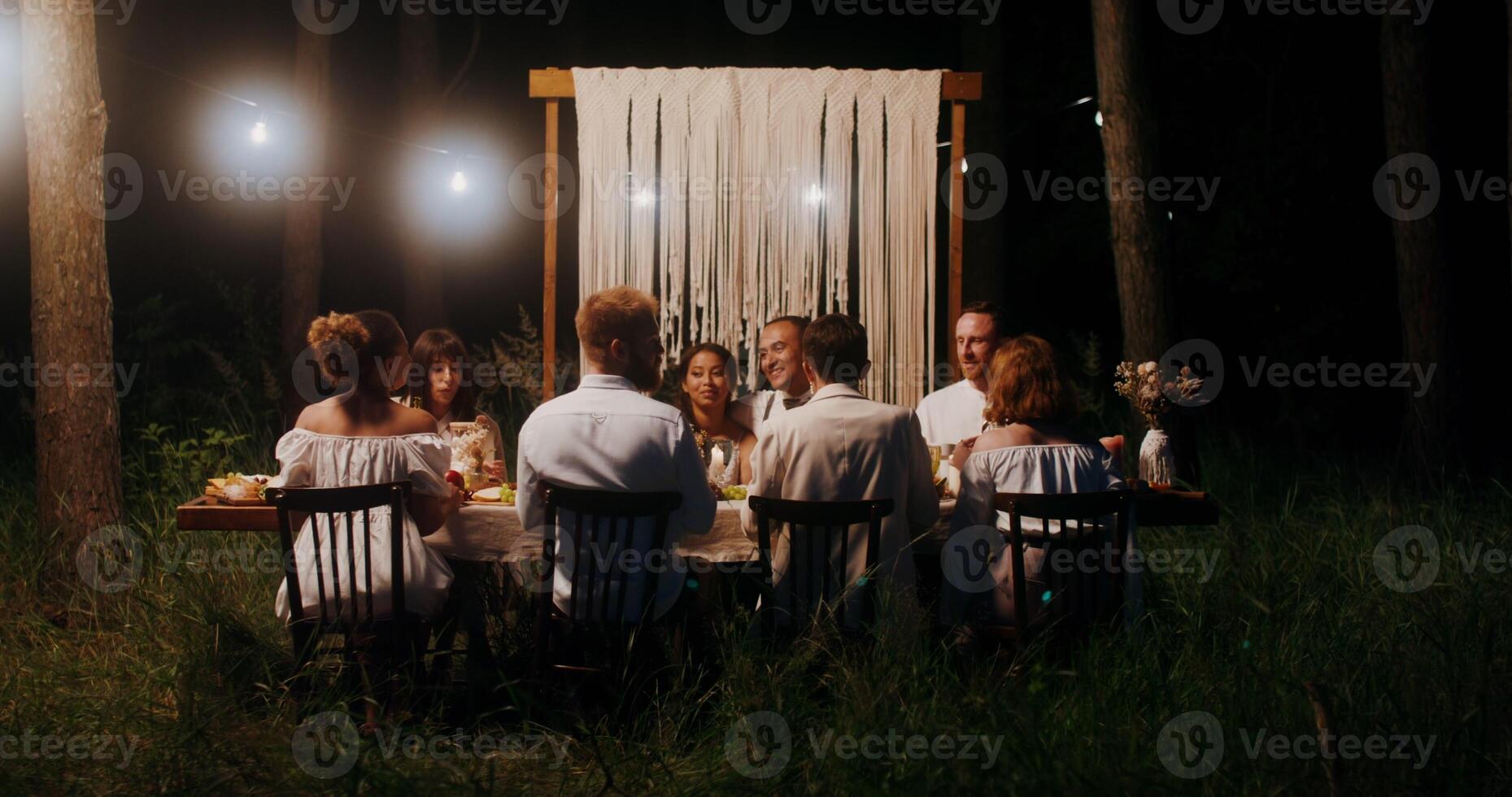 The bride and groom embrace sitting at the festive table next to their guests. A man takes pictures of the bride and groom on his mobile. Outdoor wedding feast photo
