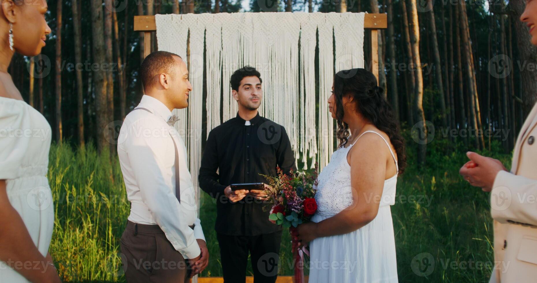 Outdoor wedding near the decorated arch with a priest and guests. The bride and groom are kissing photo