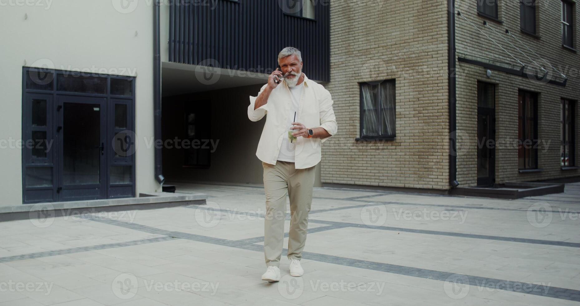 A gray-haired man with a mustache and beard talking on a mobile phone and drinking lemonade while walking along a city street - lemonade photo