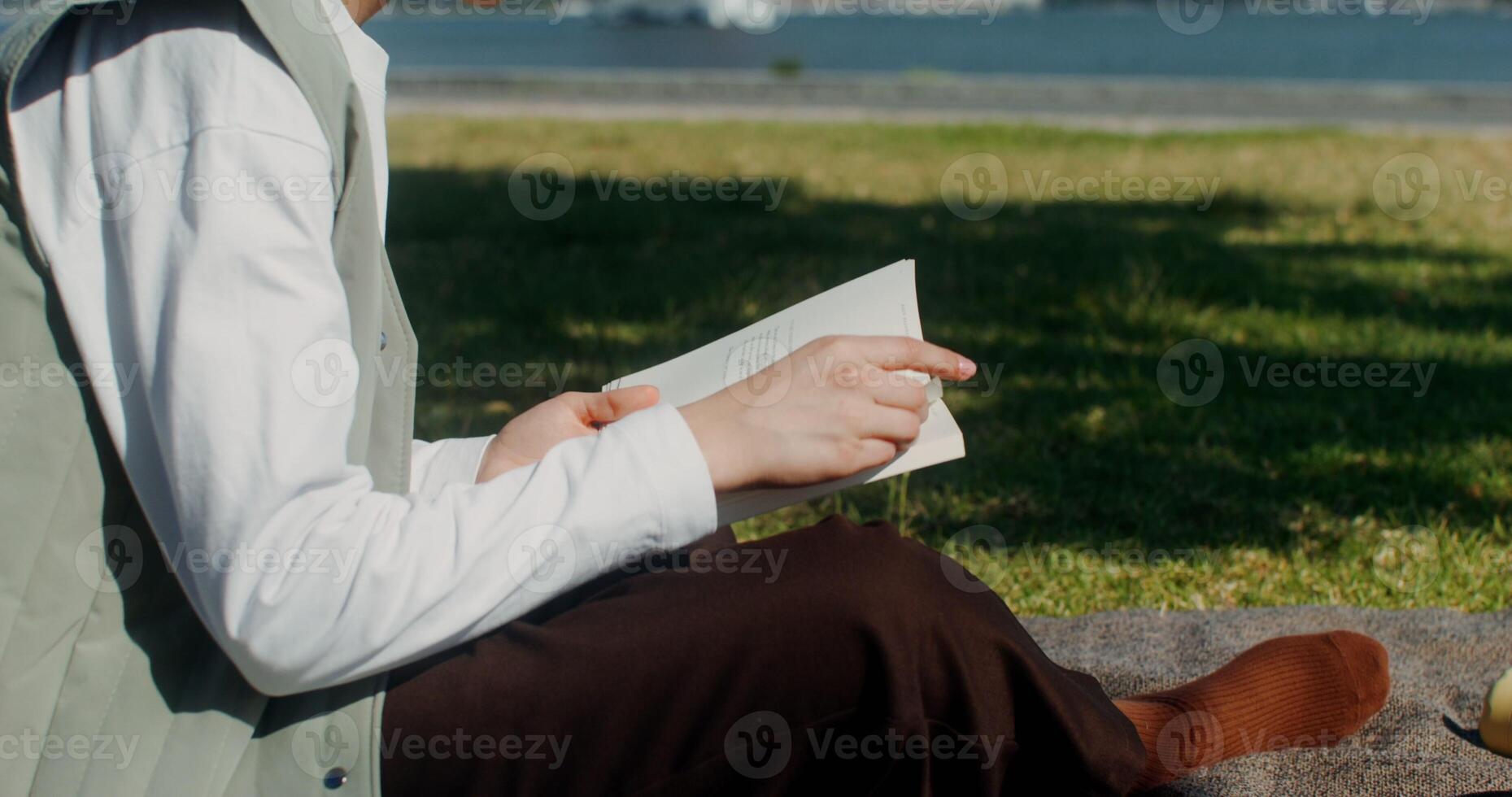 Young beautiful woman is reading a book sitting on a blanket on the riverbank near the red suspension bridge photo