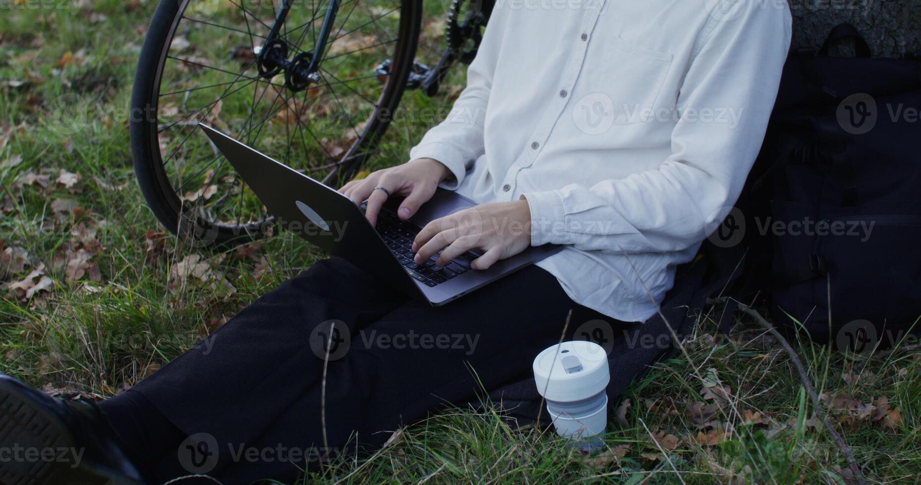 young man uses a laptop sitting on the grass under a tree outdoors. Bicycle Stands next to the man. Unidentified person, no face in the frame photo