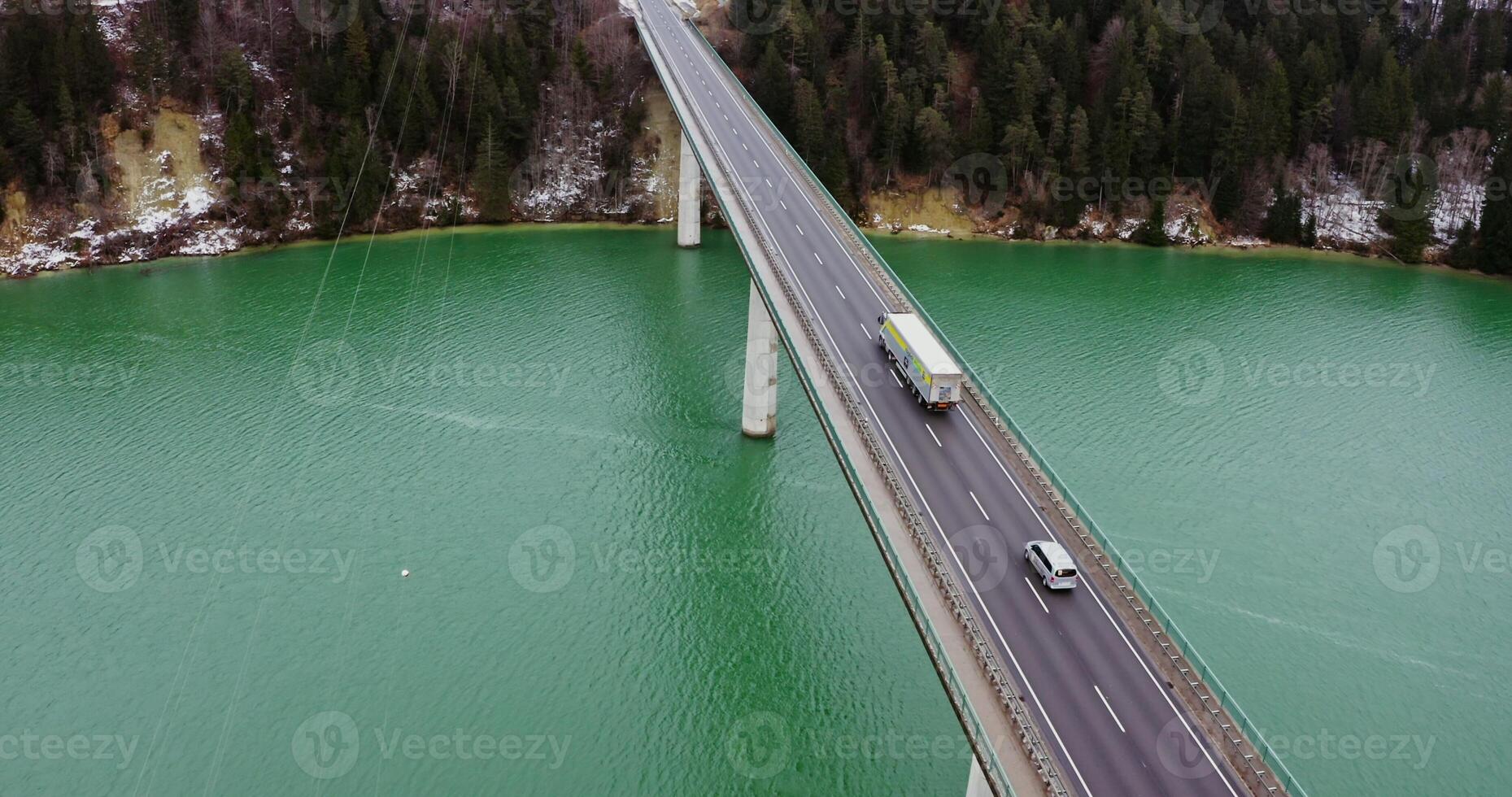 View of a concrete road bridge with a two-lane asphalt road with cars passing on it. drone footage of a long bridge across an emerald green river to hills covered in snowy forest. photo