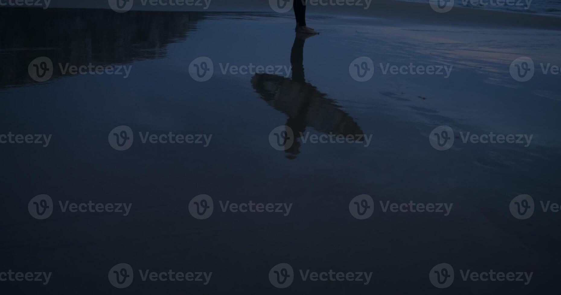 Surfer with a board in his hands stands on the seashore at dusk, reflected in the water on the shore photo