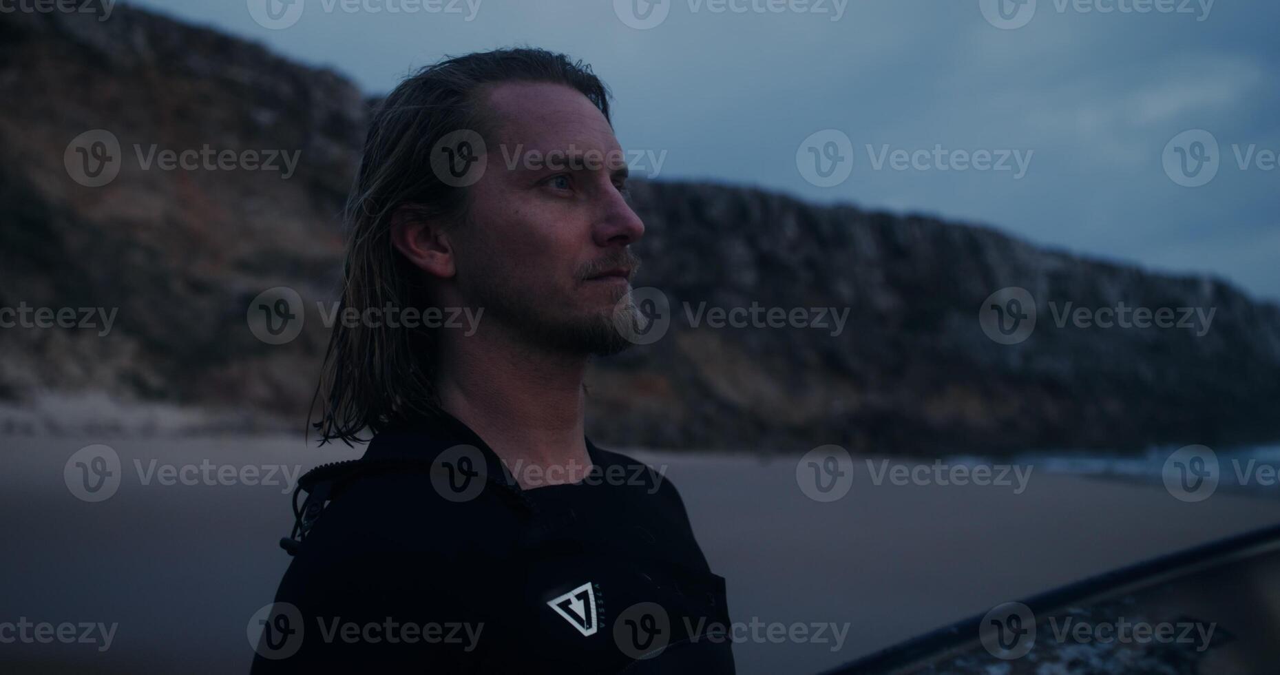 Bearded man in a diving suit, with a surfboard in his hands, smiles while looking at the camera standing on the seashore at dusk photo