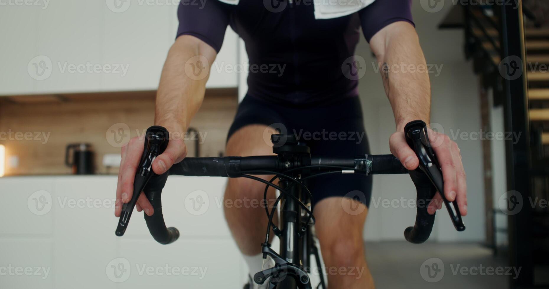 A man in headphones with a towel around his neck drinks water from a bottle, while exercising on a cycling simulator in a modern home interior photo