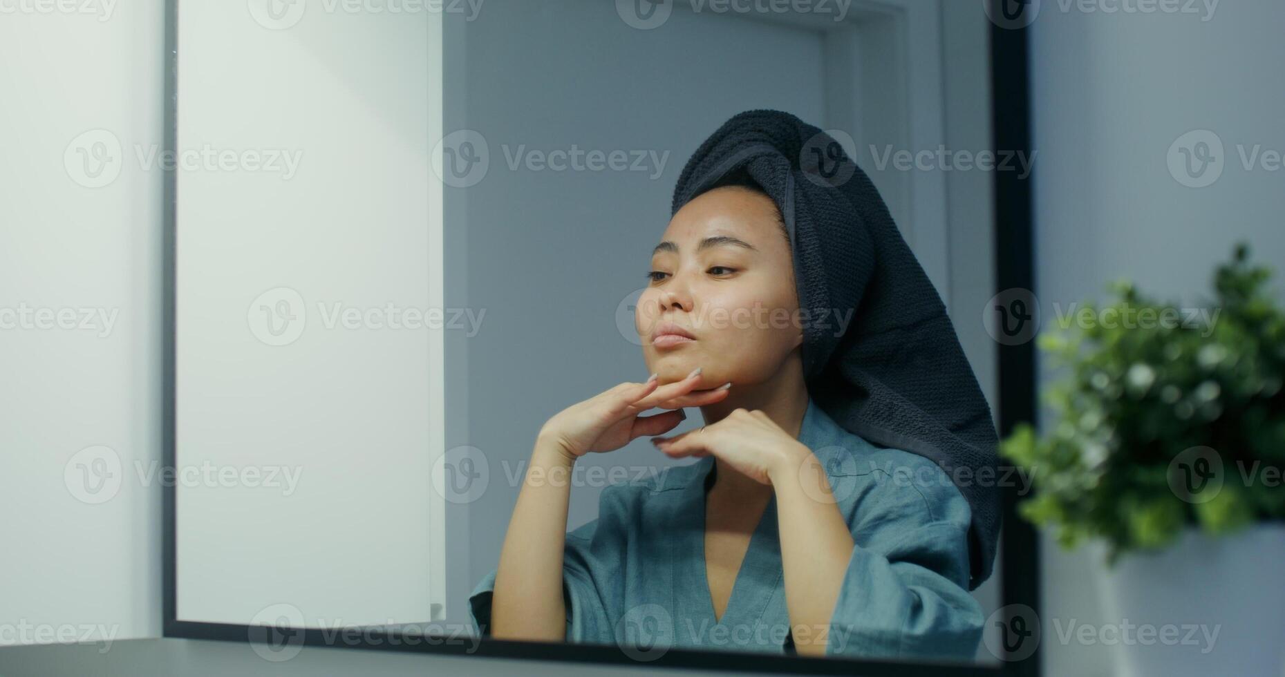 Young woman with a towel around her head is doing facial massage while looking in the mirror in a stylish bathroom photo