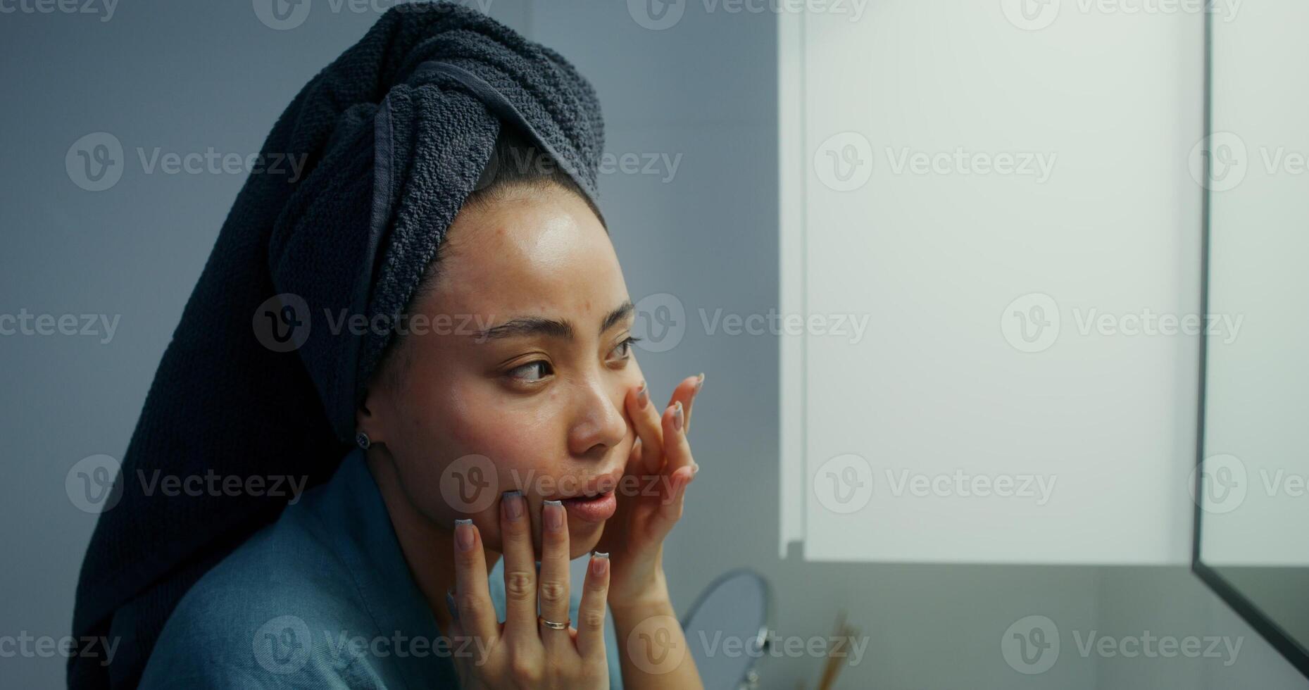 A young woman rubs a moisturizer into her face skin while looking in the mirror, her face from the side close-up photo