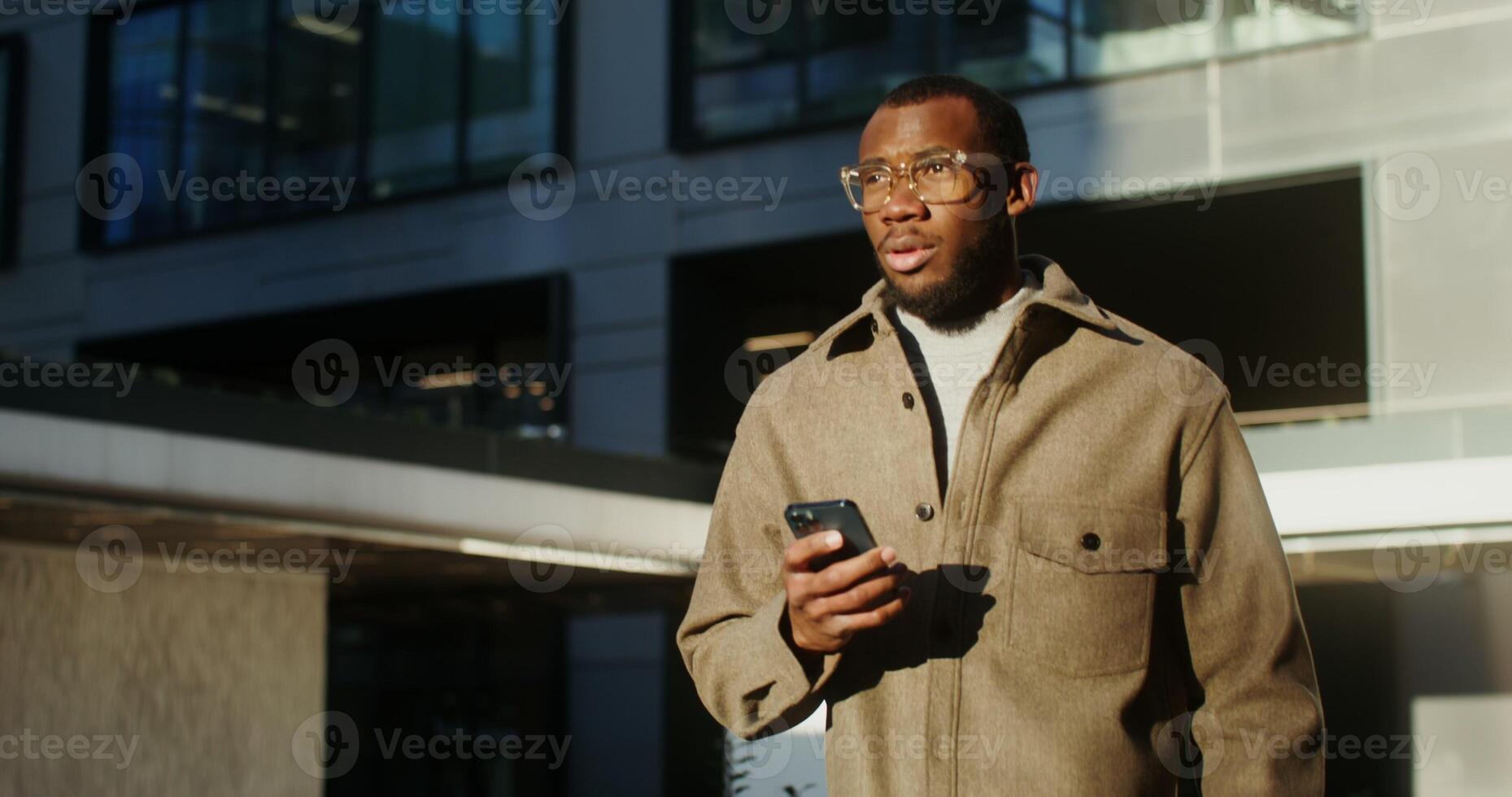 A young stylishly dressed African-American man in transparent glasses smiles while typing a text message on his mobile phone while walking near city buildings photo