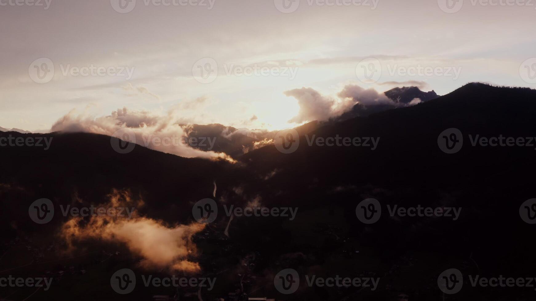 The light of rising sun breaks through fluffy white clouds over the peaks of the Alps. Village in a lowland between mountains at dusk, drone , Austria photo
