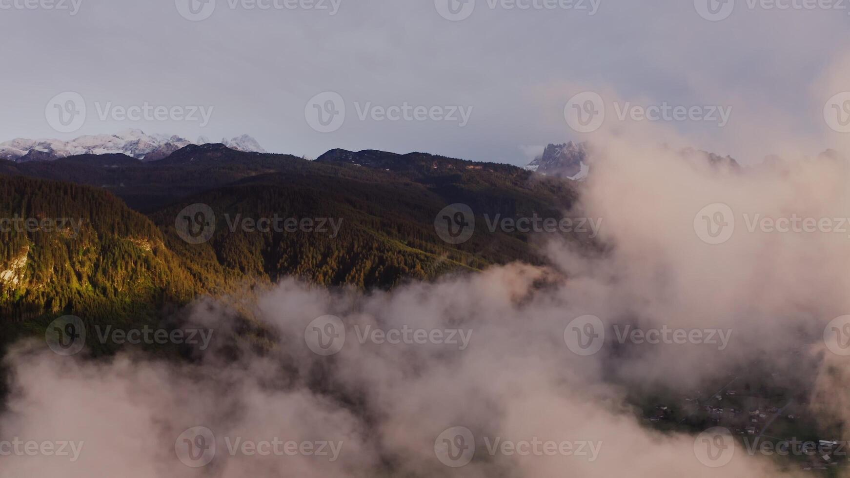 Scenic view of mountain valley from a bird's eye view through the clouds. A village in a lowland between gently sloping mountains overgrown with dense forest, drone . Austria, Alps photo