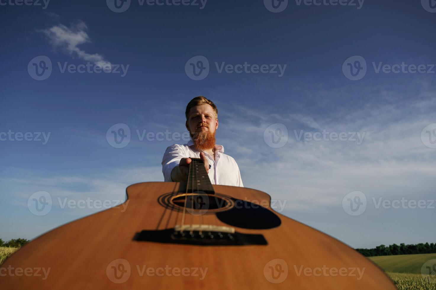 Holding acoustic guitar. Man in white shirt is on the agricultural field photo