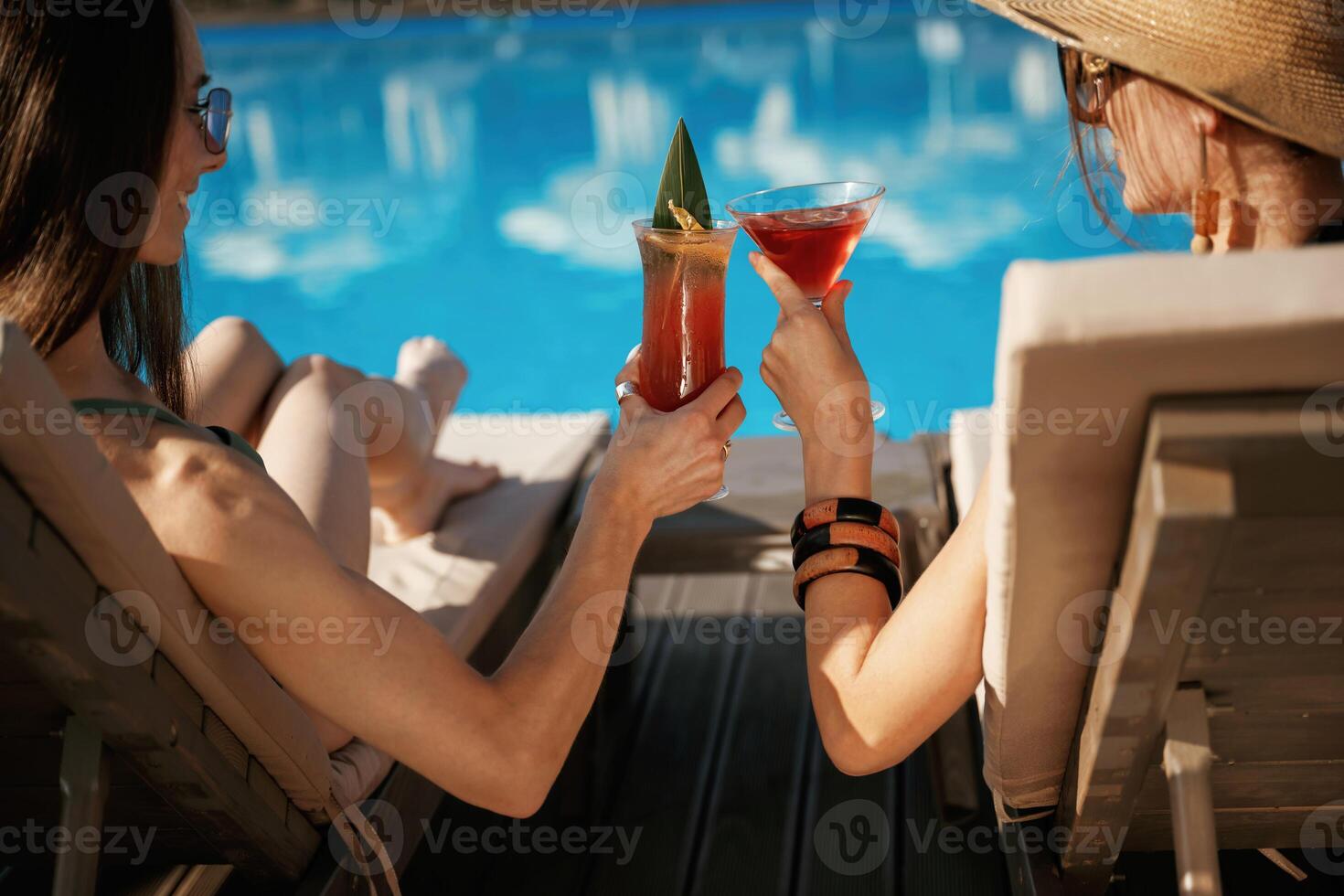 Knocking glasses, tasty drink, rear view. Two beautiful women are resting near the pool photo
