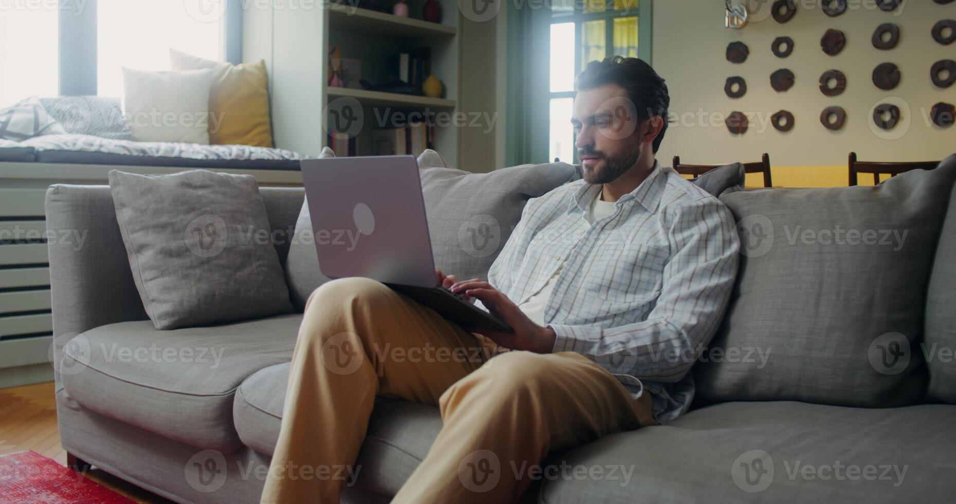 Man in casual clothes smiles while typing on a laptop while sitting on the couch at home photo