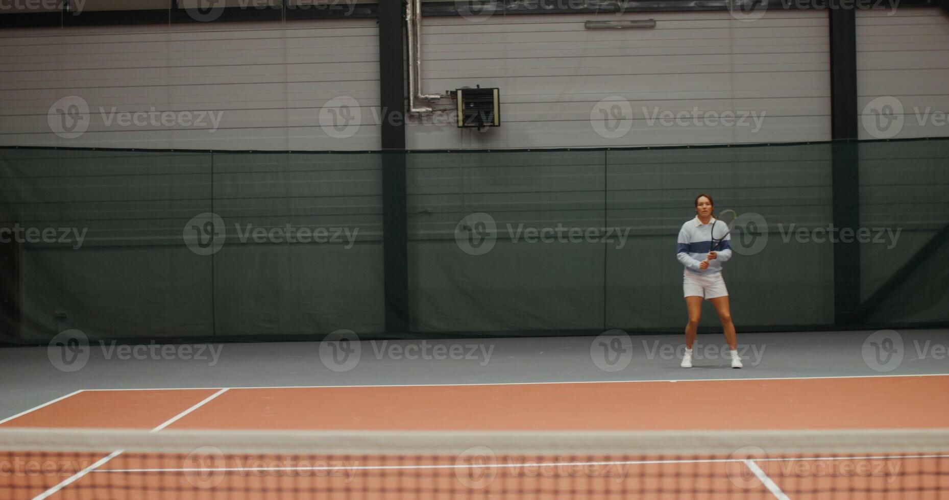 Young woman of European appearance in a sports uniform actively bouncing and hitting a tennis ball with a racket, playing tennis with a partner. Filming from a net stretched in the middle of court photo