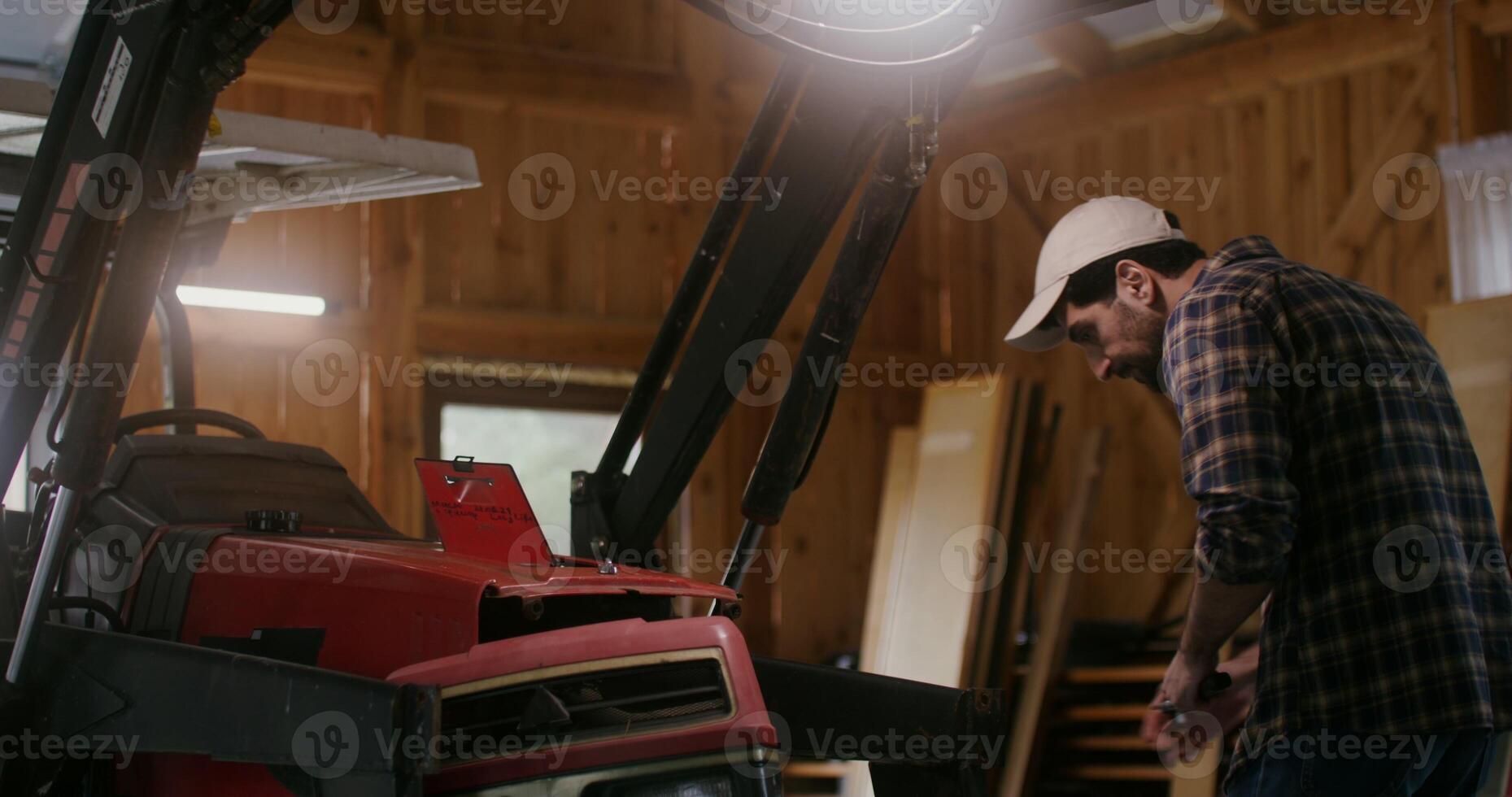 European man with a beard repairs a small agricultural machine using tools in a wooden barn, under artificial lighting photo