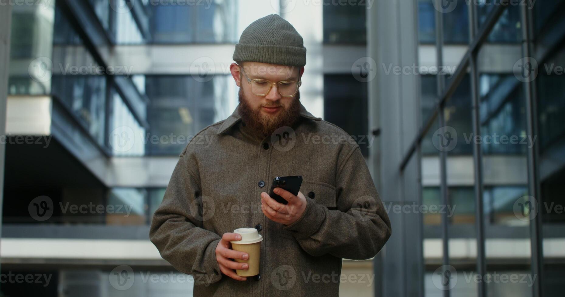 Warmly dressed bearded man in glasses is typing text on a mobile phone while standing near a stylish office building with a disposable cup in his hands. in 4k, red komodo photo