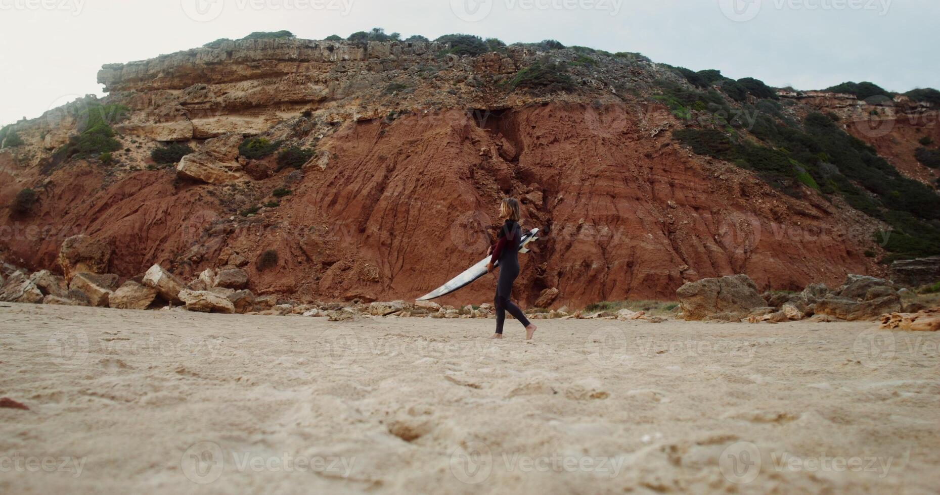 Young man in a diving suit pushes a surfboard into the sand on the beach near the red rocks photo