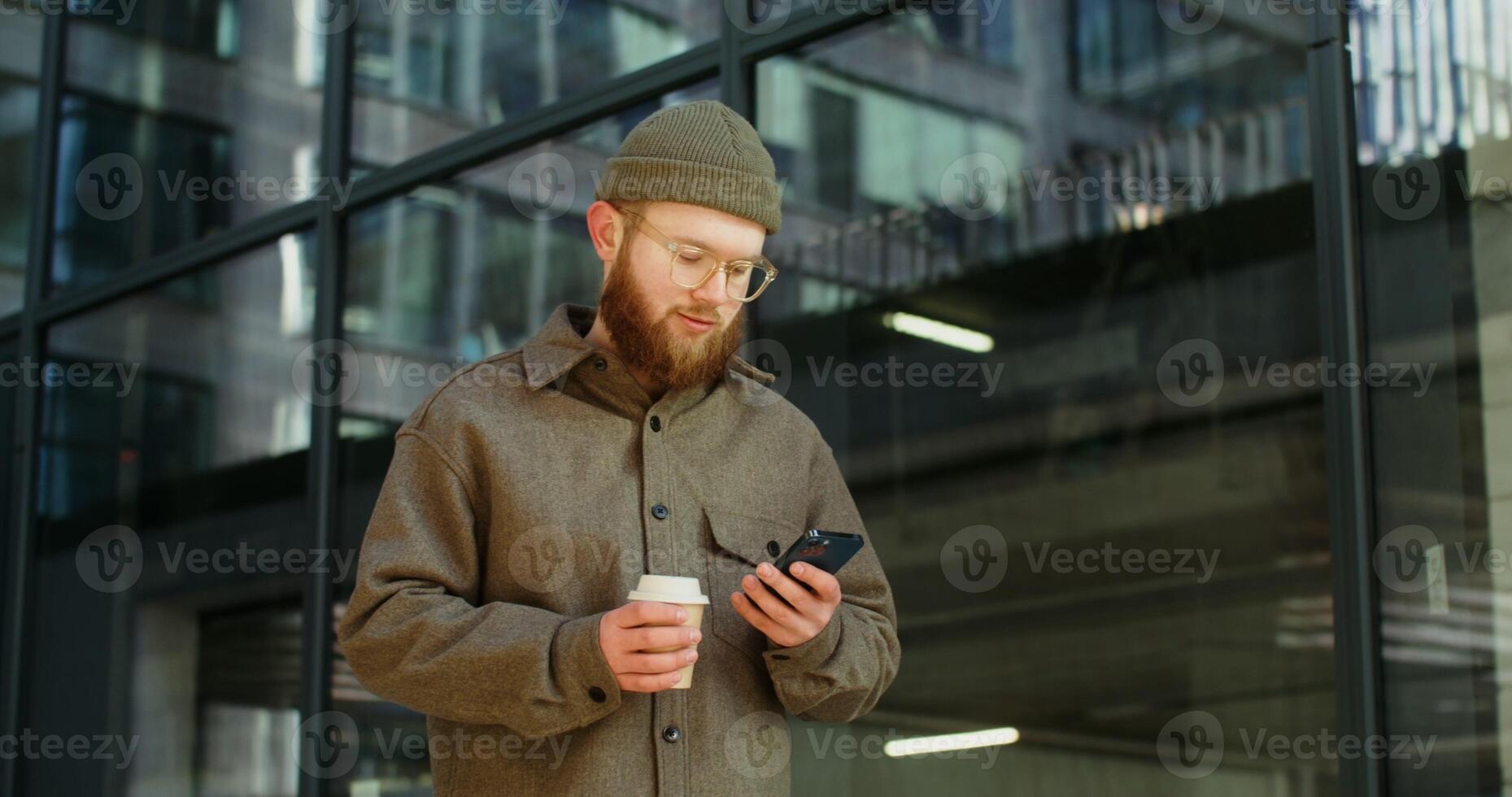 Bearded man in glasses uses a mobile phone while standing near a stylish office building with a disposable cup in his hands. Dynamic circular motion . in 4k, red komodo photo