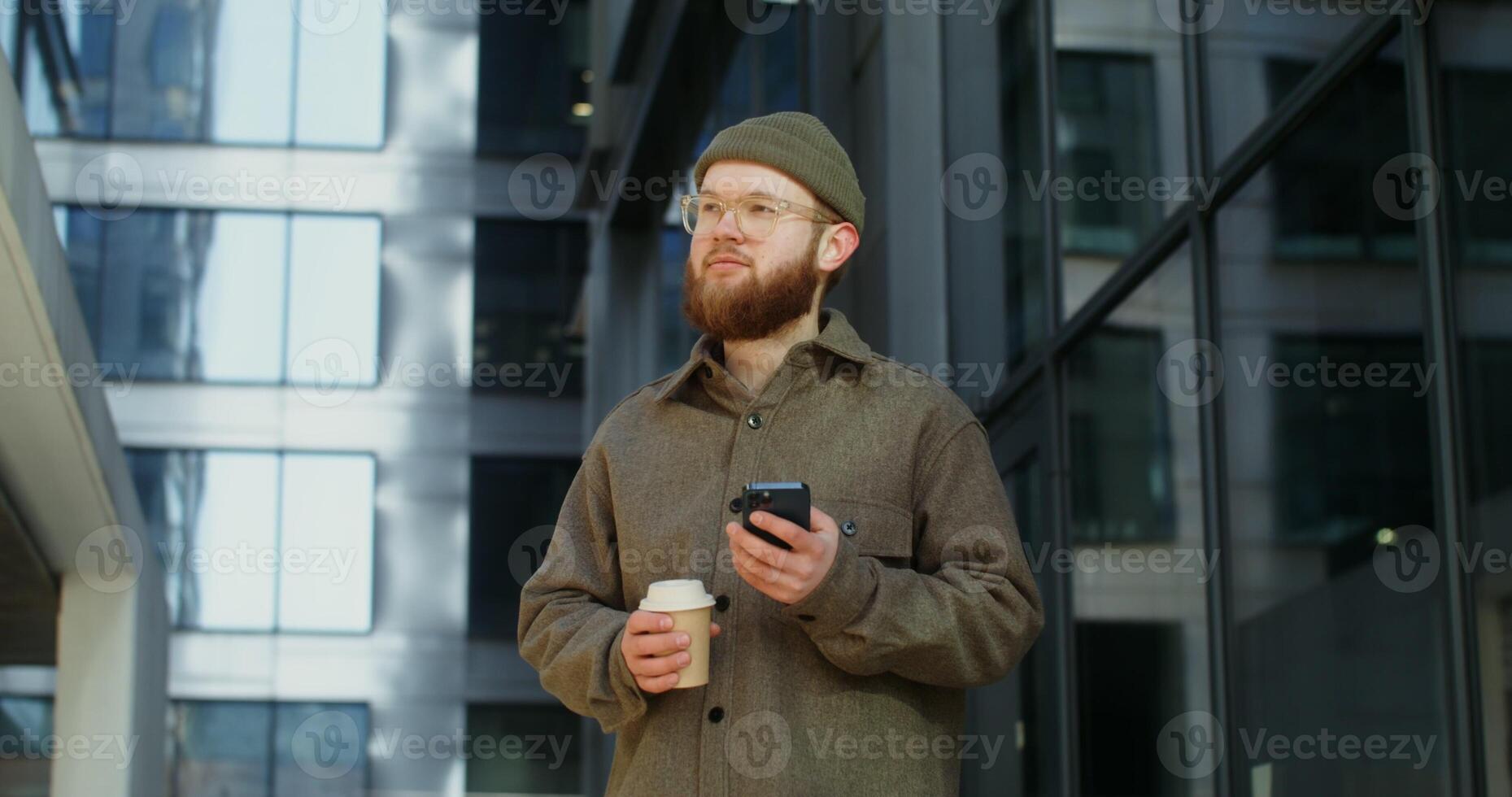 Bearded stylishly dressed man smiling while using a mobile phone while standing near an office building with large darkened windows. in 4k, red komodo photo