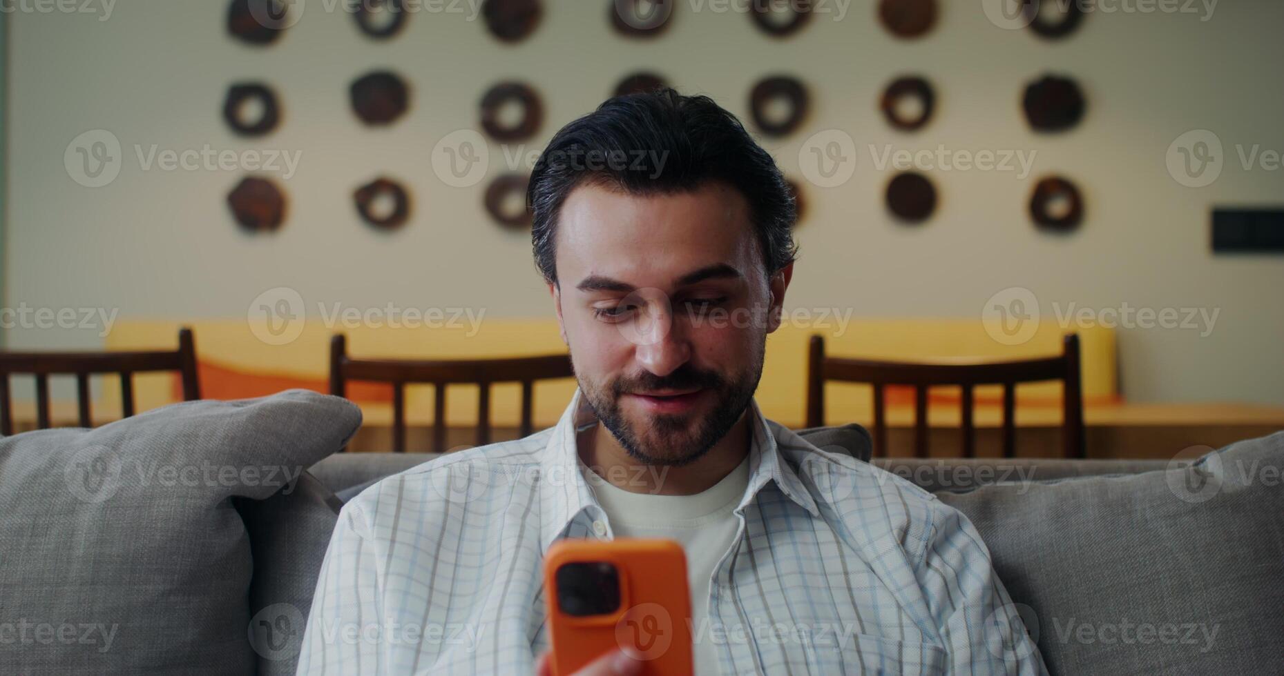 A man in casual clothes smiles and while sitting on a sofa in a stylish home interior. Front view photo