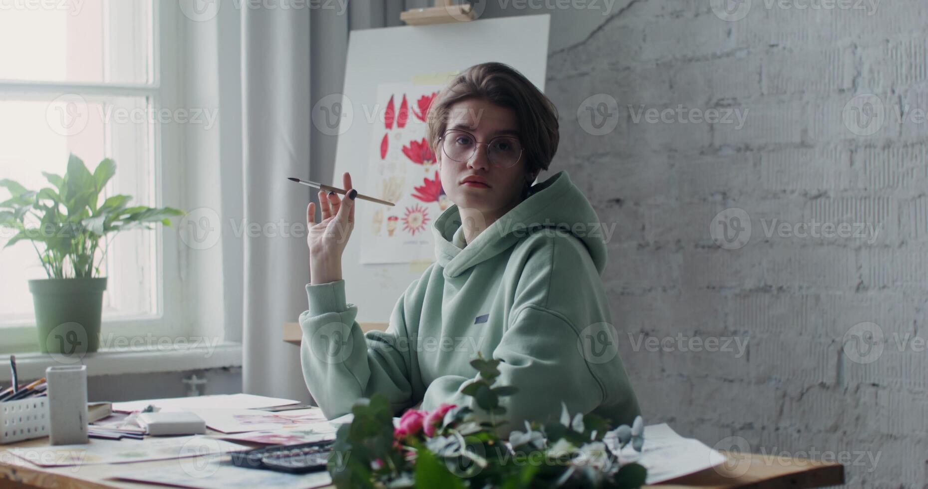 Young artist in glasses looking at the camera, while posing with a drawing brush in her hands while sitting at a desk with drawings laid out on it photo