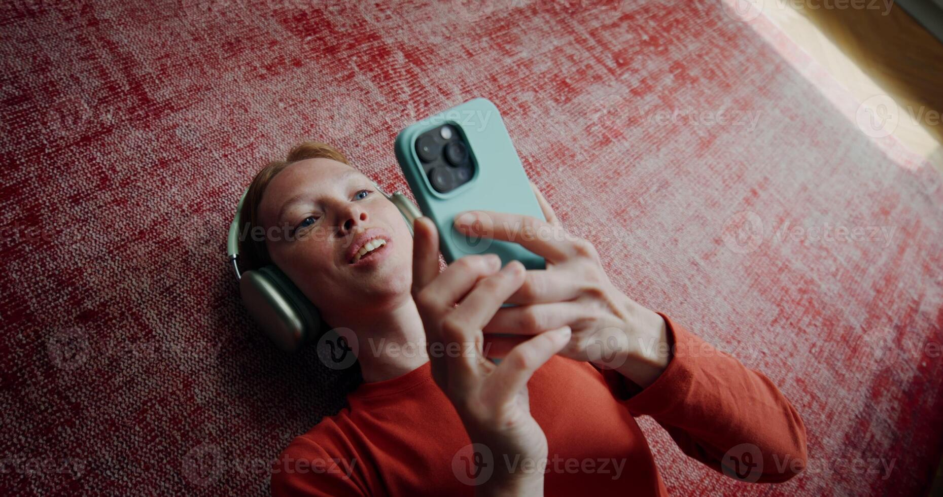 Woman in casual clothes, wearing headphones, laughing when using a mobile phone while she lying on the floor at home. The moves from the woman's face to the general plan photo