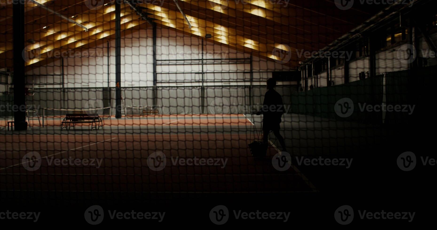 Light comes on in the gym and a young man of European appearance walks to the bench, carrying a racket and a basket full of tennis balls, getting ready to play tennis. Filming through a grid photo
