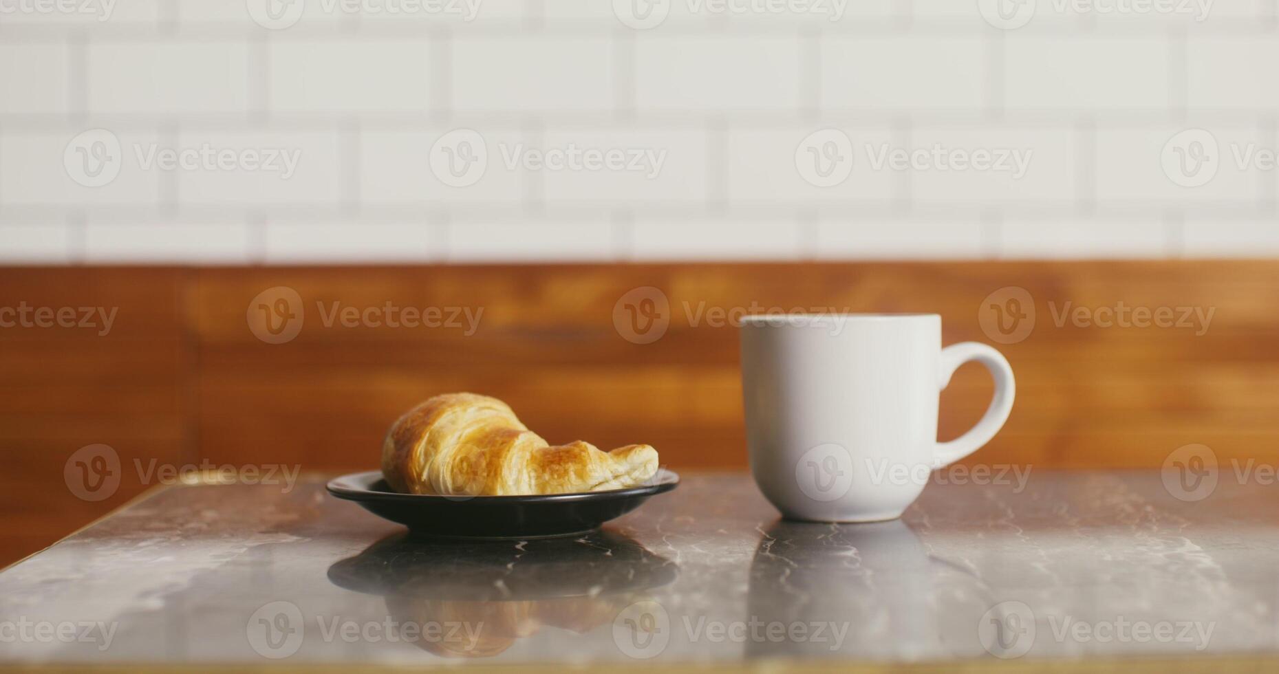 Large white mug and a black plate with a croissant stand on a small coffee table, prepared for a regular customer. No people on photo