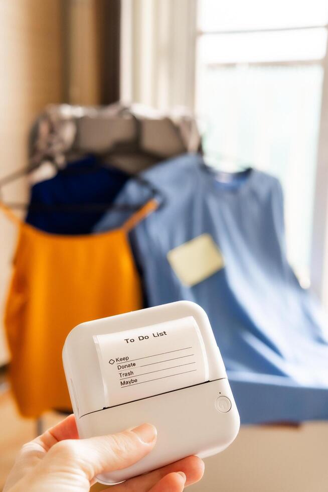 Hand holding mini thermal printer with clothing decluttering checklist in bright minimalist room with organized garments on hangers in background concept of smart home wardrobe planning and sorting photo