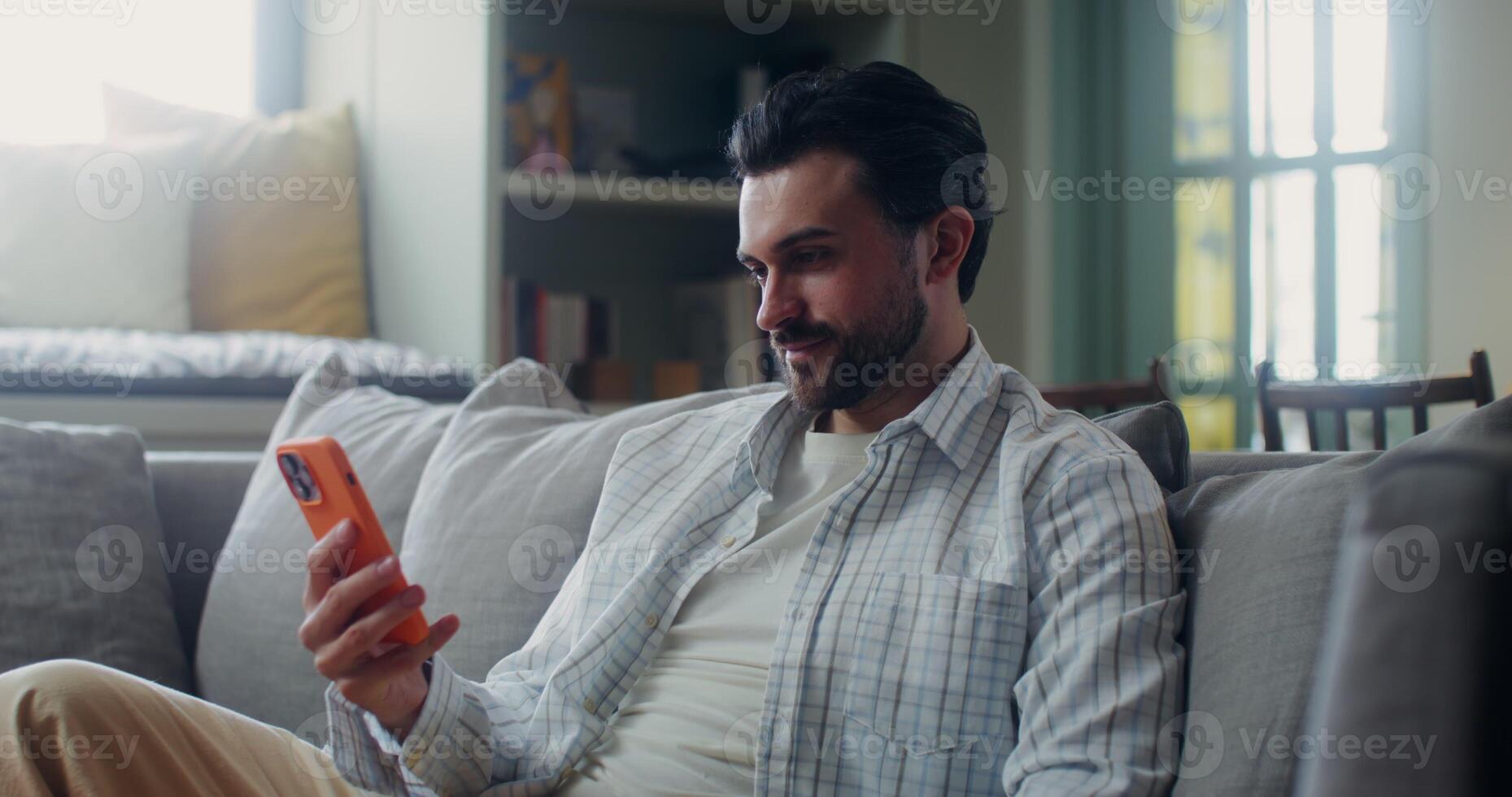 Man in casual clothes smiles, and waves hello starting a call on a mobile phone while sitting on a sofa in a stylish home interior photo