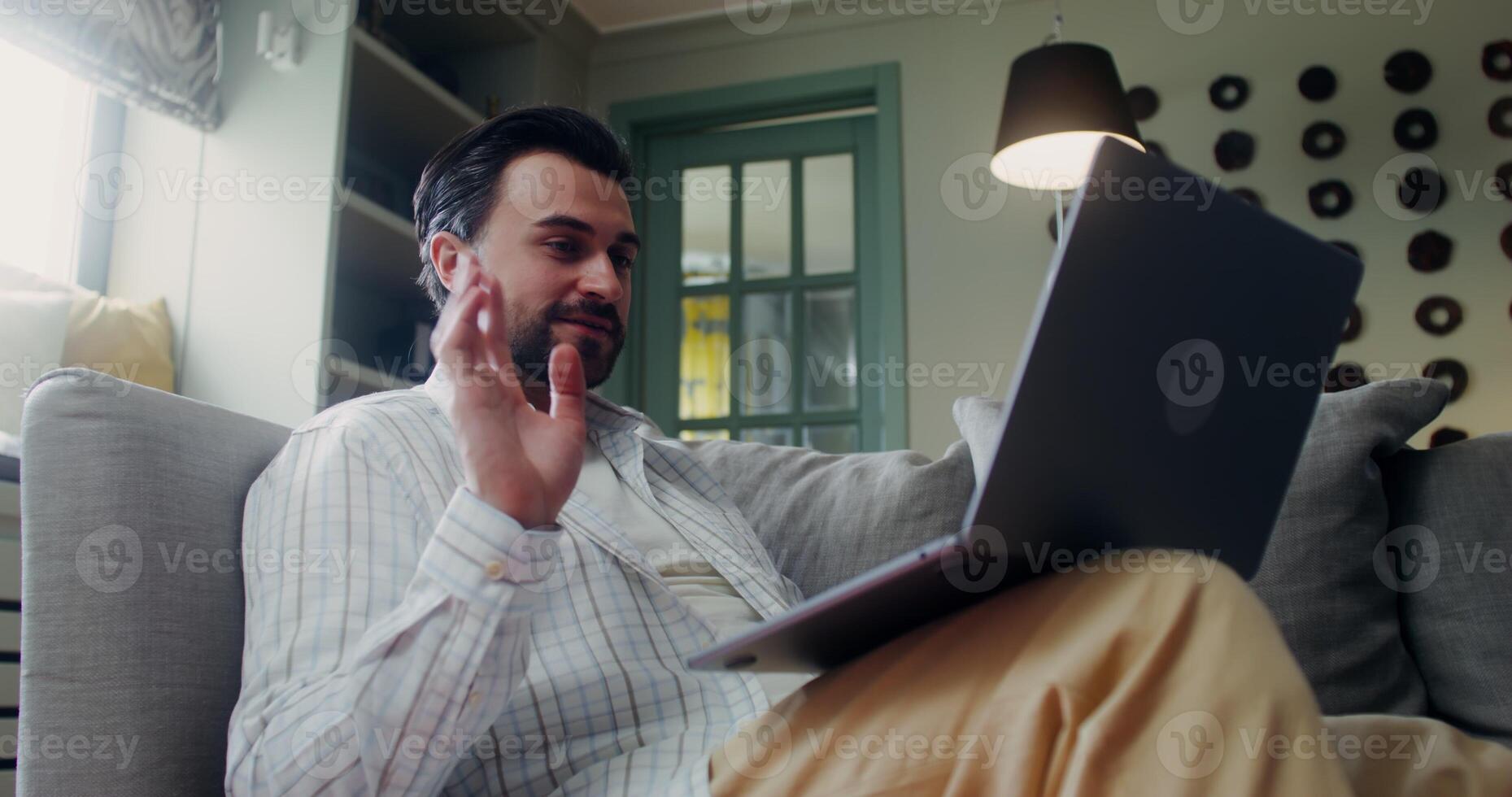 A stylish bearded man smiles and waves hello on a laptop standing on his lap, sitting on a sofa indoors photo